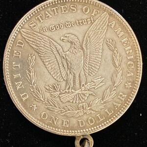 May include: Silver dollar coin with a bald eagle and the words "United States of America" and "In God We Trust" on the front. The coin has a loop on the bottom for hanging.