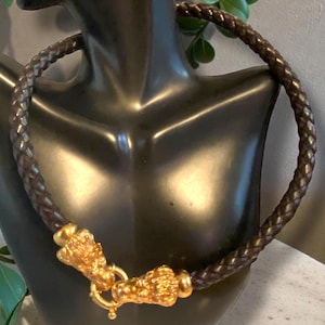May include: Braided brown leather necklace with gold-colored lion head clasp. The necklace is displayed on a black mannequin. The clasp features intricate details and a secure closure. The leather appears to be tightly woven.