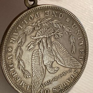 May include: Silver dollar coin with a bald eagle perched on a branch with its wings spread. The coin has the words "One Dollar", "United States of America", and "In God We Trust" inscribed around the eagle.