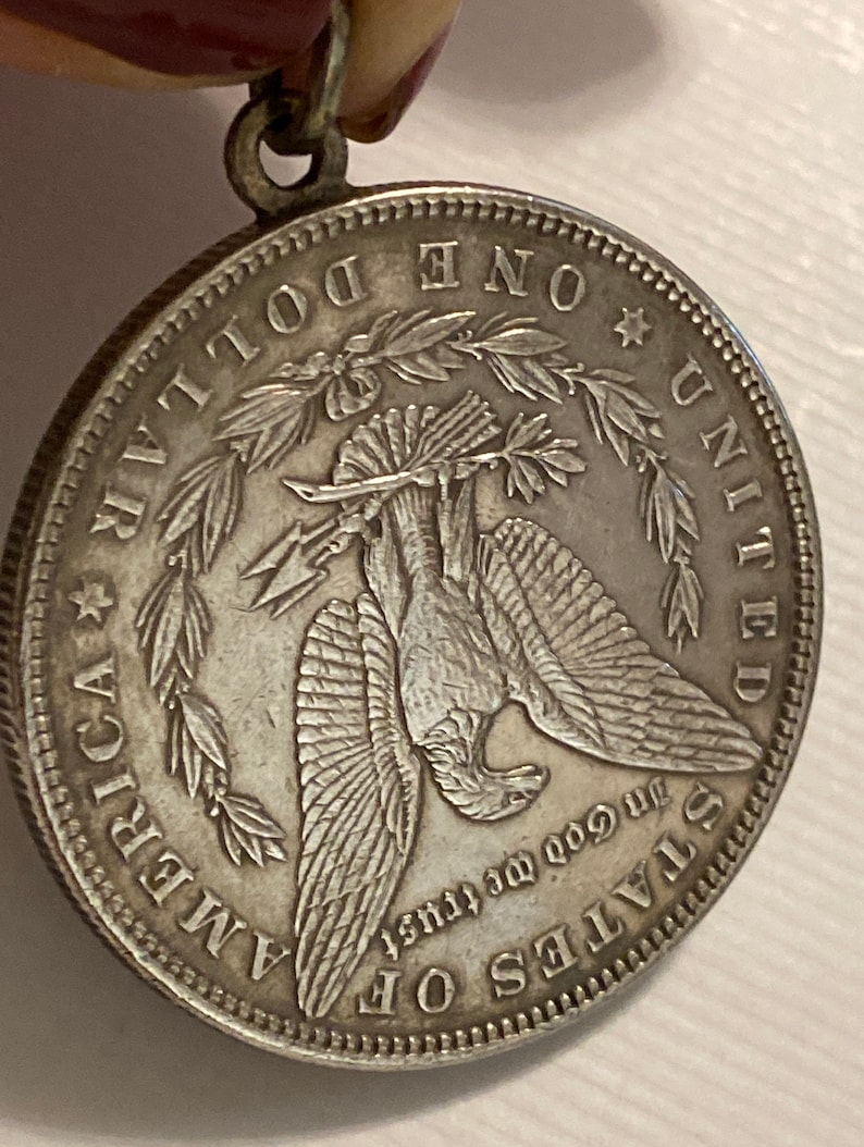 May include: Silver dollar coin with a bald eagle perched on a branch with its wings spread. The coin has the words "One Dollar", "United States of America", and "In God We Trust" inscribed around the eagle.