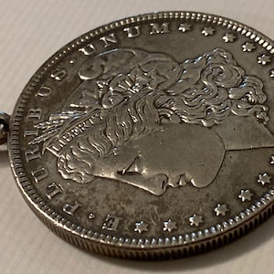 May include: A silver Morgan dollar coin with a loop attached to the top. The coin features a profile of Lady Liberty with the inscription "LIBERTY" and the date "1883".