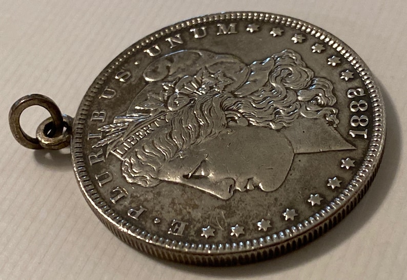 May include: A silver Morgan dollar coin with a loop attached to the top. The coin features a profile of Lady Liberty with the inscription "LIBERTY" and the date "1883".