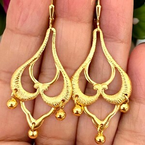 May include: A pair of gold dangle earrings with a delicate, intricate design. The earrings feature multiple layers of openwork hearts and a dangling ball at the bottom.