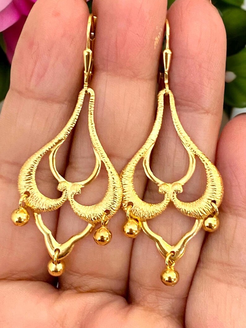 May include: A pair of gold dangle earrings with a delicate, intricate design. The earrings feature multiple layers of openwork hearts and a dangling ball at the bottom.