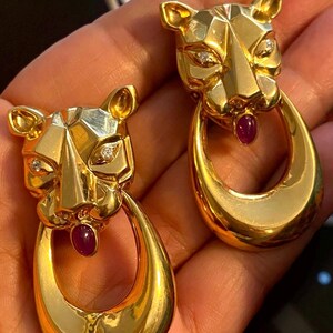 May include: A pair of gold-toned earrings featuring a stylized panther design. Each earring has a faceted head with diamond accents and a teardrop-shaped loop. A small, red gemstone is set in the mouth of each panther.