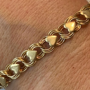 May include: A gold-toned bracelet featuring a series of heart-shaped charms linked together. The bracelet's design incorporates double-linked circular segments, creating a chain-like appearance. The hearts are evenly spaced along the length of the bracelet.