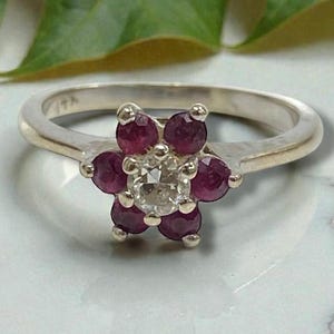 May include: A silver ring featuring a flower design. The flower's petals are made of deep red gemstones, surrounding a central clear gemstone. The ring is set on a light-colored surface, with a green leaf in the background.