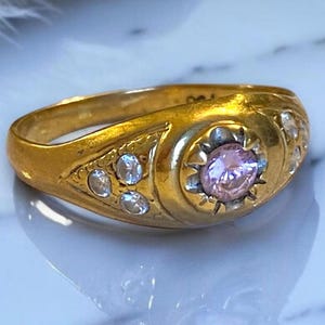 Pu&ograve; includere: Un anello d'oro con una pietra preziosa rosa circondata da pietre preziose pi&ugrave; piccole e trasparenti. L'anello ha un design dettagliato e una fascia arrotondata.
