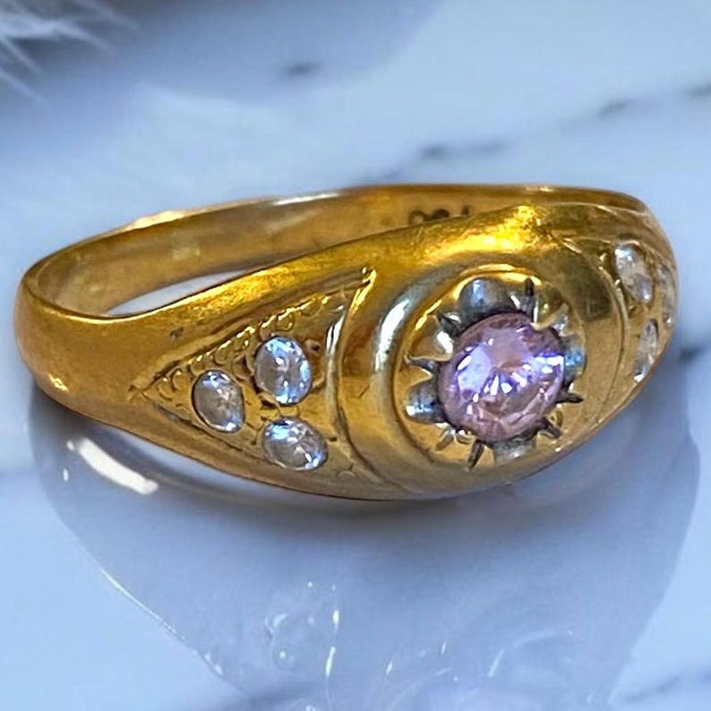 Pu&ograve; includere: Un anello d'oro con una pietra preziosa rosa circondata da pietre preziose pi&ugrave; piccole e trasparenti. L'anello ha un design dettagliato e una fascia arrotondata.