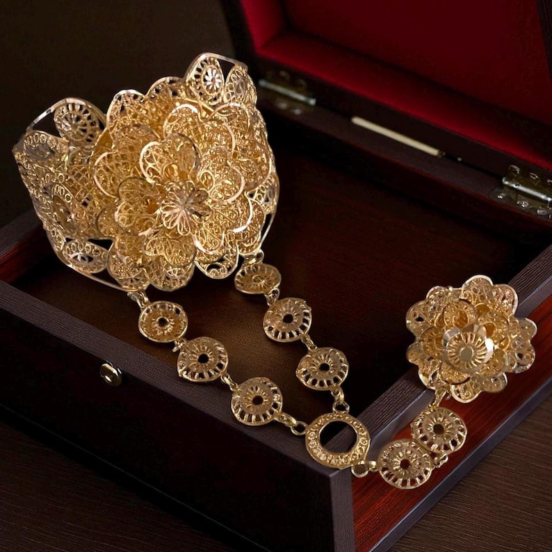 May include: A gold-colored filigree jewelry set featuring a large floral cuff bracelet, a ring, and a chain of circular links. The jewelry is displayed inside a dark wooden box with a red interior.