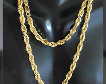 May include: A gold-colored rope chain necklace displayed on a black mannequin. The necklace is made of twisted links, creating a rope-like appearance. The chain is draped in a layered style, showcasing its design and texture. The clasp is visible.