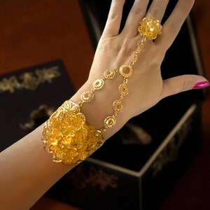 May include: A gold-colored hand chain bracelet with a floral design. The bracelet connects to a ring with a matching flower. The bracelet features a large floral cuff and a series of circular links. The background includes a black box.