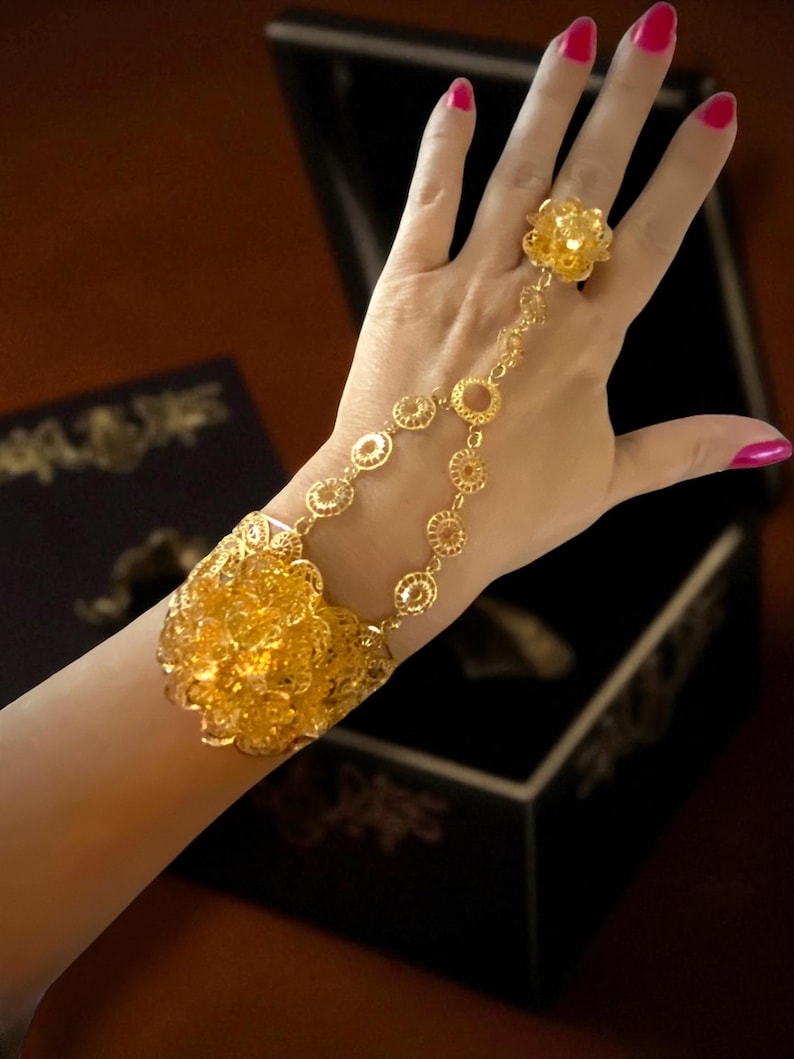 May include: A gold-colored hand chain bracelet with a floral design. The bracelet connects to a ring with a matching flower. The bracelet features a large floral cuff and a series of circular links. The background includes a black box.