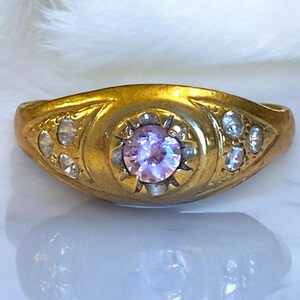 Pu&ograve; includere: Un anello d'oro con una singola pietra preziosa rotonda di colore viola chiaro circondata da pietre preziose pi&ugrave; piccole e chiare. L'anello ha un design semplice ed elegante.
