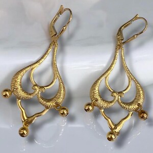 May include: A pair of gold dangle earrings with a delicate, intricate design. The earrings feature a heart-shaped motif with multiple loops and small round beads hanging from the bottom.