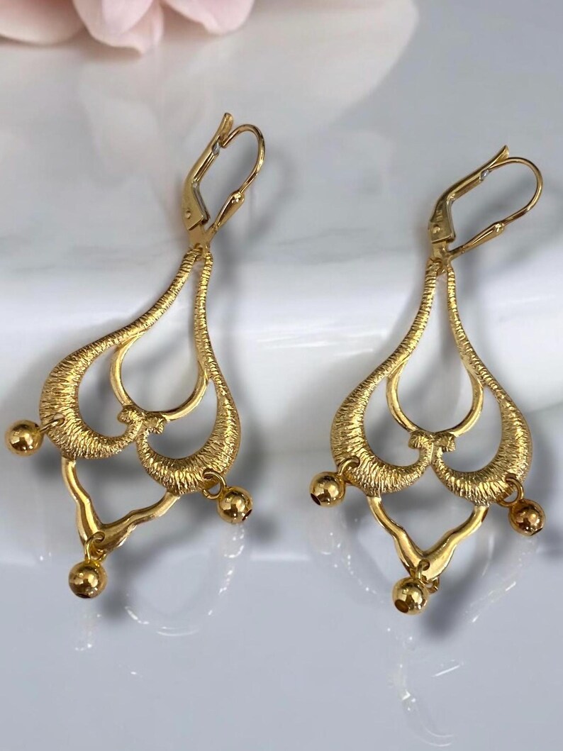 May include: A pair of gold dangle earrings with a delicate, intricate design. The earrings feature a heart-shaped motif with multiple loops and small round beads hanging from the bottom.
