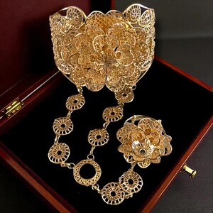 May include: A gold-colored filigree jewelry set, including a wide cuff bracelet, a ring, and a chain of circular links. The pieces are displayed inside a wooden box with a dark interior. The jewelry has an intricate floral design.