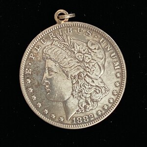 May include: A silver US Morgan dollar coin with a portrait of Lady Liberty on the obverse. The coin is dated 1882 and has the inscription "E Pluribus Unum" around the edge.