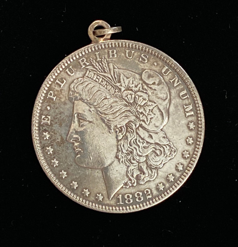 May include: A silver US Morgan dollar coin with a portrait of Lady Liberty on the obverse. The coin is dated 1882 and has the inscription "E Pluribus Unum" around the edge.