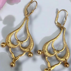 May include: A pair of gold dangle earrings with a teardrop shape and multiple loops. The earrings have a textured surface and are accented with small gold balls.