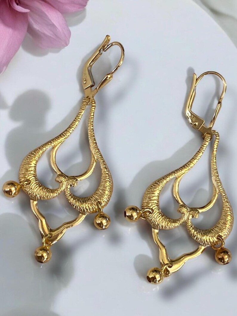 May include: A pair of gold dangle earrings with a teardrop shape and multiple loops. The earrings have a textured surface and are accented with small gold balls.
