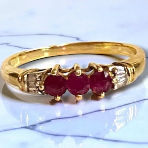 May include: A gold ring featuring three vibrant red gemstones set in a row. The ring has a classic design with a polished gold band and small accent stones on either side of the gemstones. The gemstones are a deep red color.