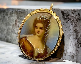 May include: A gold locket with a portrait of a woman in a brown dress. The woman has a white lace collar and her hair is styled in a bun with a white ribbon. The locket has a gold chain.