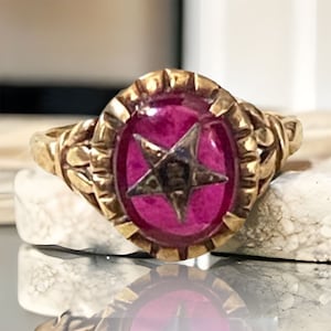 May include: A gold ring with a pink oval stone set in a decorative frame. The stone has a five-pointed star design in the center.