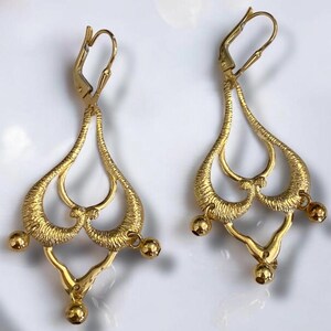 May include: A pair of gold dangle earrings with a delicate, intricate design. The earrings feature a teardrop shape with multiple loops and small round beads hanging from the bottom.