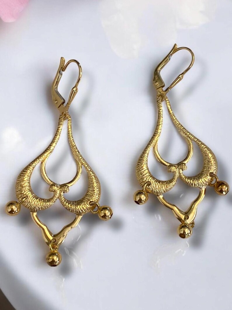 May include: A pair of gold dangle earrings with a delicate, intricate design. The earrings feature a teardrop shape with multiple loops and small round beads hanging from the bottom.