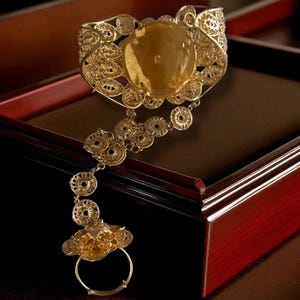 May include: Ornate gold-toned hand bracelet with filigree detailing. The bracelet features a large, oval-shaped central stone and a delicate chain connecting to a ring. The design includes circular and floral patterns, creating an elegant, vintage aesthetic.