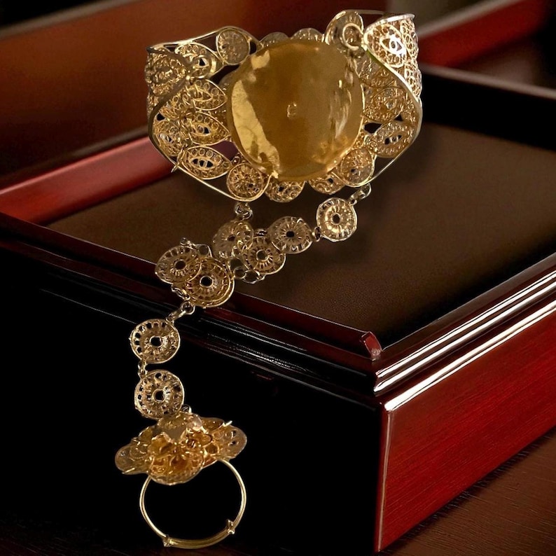 May include: Ornate gold-toned hand bracelet with filigree detailing. The bracelet features a large, oval-shaped central stone and a delicate chain connecting to a ring. The design includes circular and floral patterns, creating an elegant, vintage aesthetic.