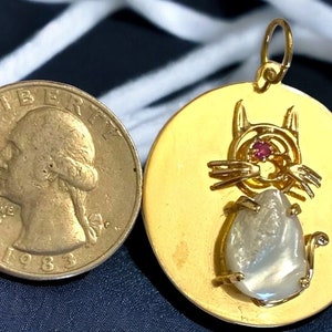 May include: A gold-colored pendant featuring a cat design with a red gemstone eye and a pear-shaped, light-colored stone body. The pendant is next to a 1983 quarter coin for size comparison. The pendant has a loop for a chain.