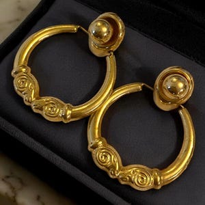 May include: A pair of gold-toned hoop earrings with a decorative swirl design. The earrings have a post and a round ball at the top. The hoops are thick and have a vintage look.