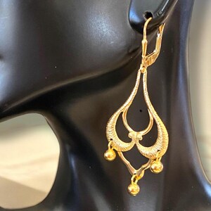 May include: Gold dangle earrings with a delicate, intricate design. The earrings feature three small, round beads hanging from the bottom.