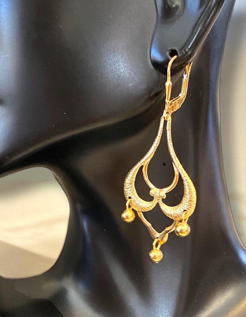 May include: Gold dangle earrings with a delicate, intricate design. The earrings feature three small, round beads hanging from the bottom.