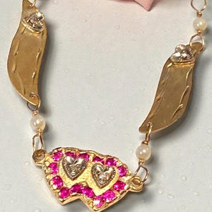 May include: A gold-toned necklace featuring a design with two heart-shaped pendants encrusted with pink gemstones. The necklace includes gold leaf-shaped elements and small pearl accents, creating a vintage aesthetic.