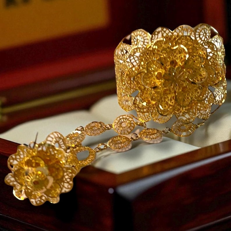 May include: A gold-colored filigree bracelet and matching necklace with floral and circular designs. The jewelry is displayed inside a wooden box with a red interior, resting on a white surface.