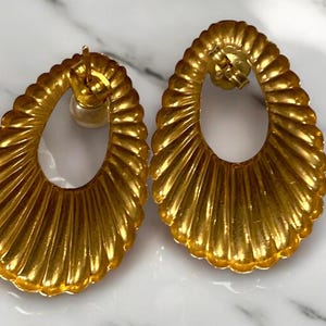 Pu&ograve; includere: Un paio di orecchini ovali in oro con un design testurizzato e nervato. Gli orecchini hanno un grande cerchio aperto al centro.