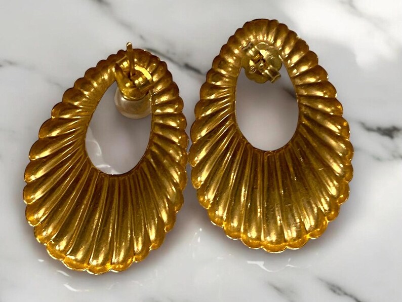 Pu&ograve; includere: Un paio di orecchini ovali in oro con un design testurizzato e nervato. Gli orecchini hanno un grande cerchio aperto al centro.