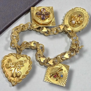 May include: A gold charm bracelet with heart-shaped links and several charms. Charms include a heart with "Sweet 16", a square with a four-leaf clover, and a round charm with "Birthday". The bracelet has a clasp.