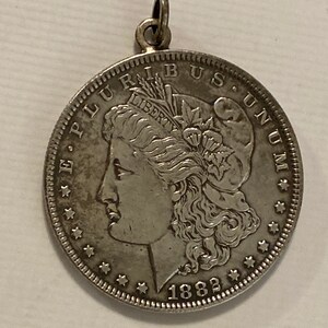 May include: Silver dollar coin pendant with a portrait of Lady Liberty on the obverse, the inscription "E PLURIBUS UNUM" and the date "1882".