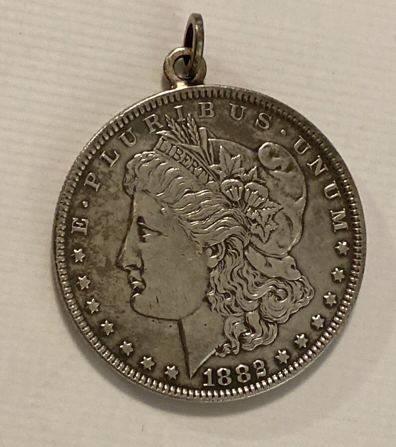 May include: Silver dollar coin pendant with a portrait of Lady Liberty on the obverse, the inscription "E PLURIBUS UNUM" and the date "1882".