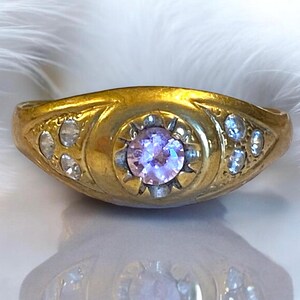 Pu&ograve; includere: Un anello d'oro con una singola pietra preziosa rotonda di colore viola chiaro circondata da pietre preziose pi&ugrave; piccole e chiare. L'anello ha un design semplice ed elegante.