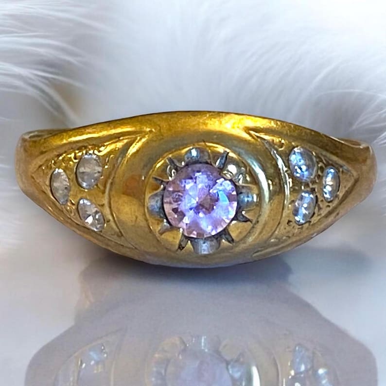 Pu&ograve; includere: Un anello d'oro con una singola pietra preziosa rotonda di colore viola chiaro circondata da pietre preziose pi&ugrave; piccole e chiare. L'anello ha un design semplice ed elegante.