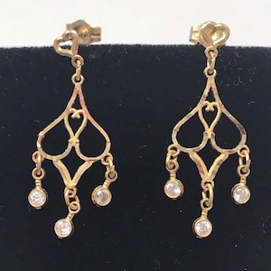 May include: A pair of gold dangle earrings with a delicate, intricate design. Each earring features a heart-shaped pendant with three small, clear stones hanging from it.