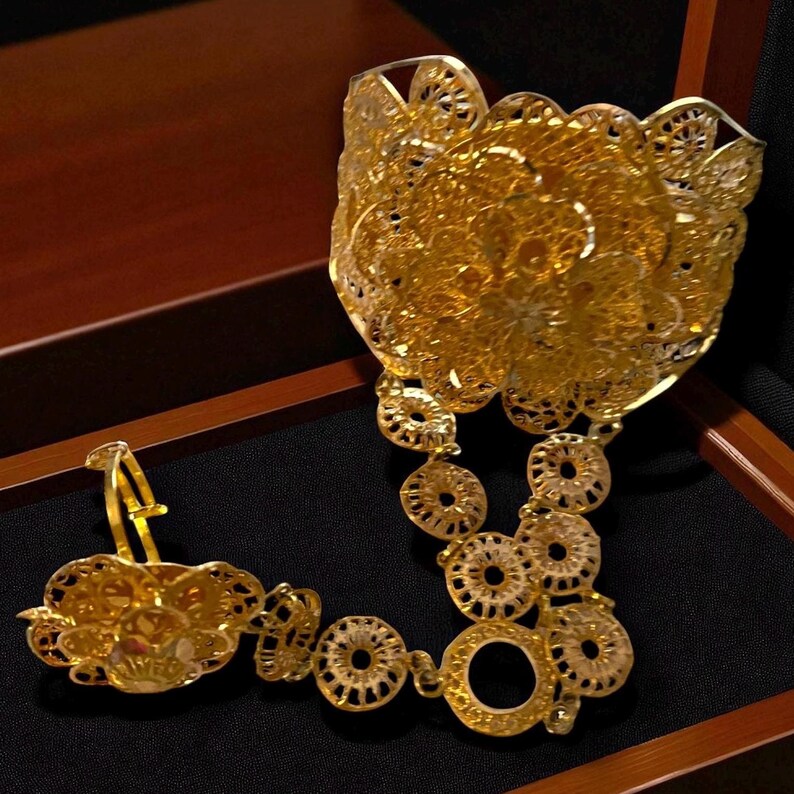 May include: Ornate gold-colored jewelry set featuring a filigree design. The set includes a large cuff bracelet, a necklace with circular links, and a ring. The pieces are displayed on a black surface within a wooden box.