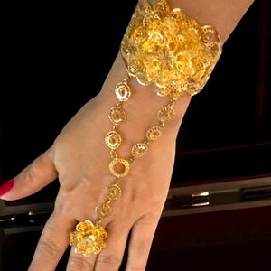 May include: A gold-colored hand chain bracelet featuring a large, ornate cuff and a delicate chain connecting to a floral ring. The jewelry has intricate filigree details and a bright, polished finish.