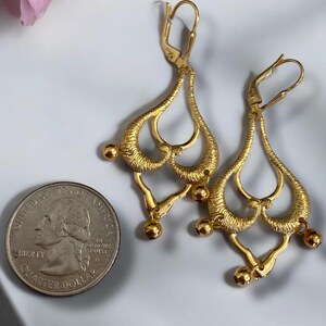 May include: A pair of gold-tone dangle earrings with a teardrop shape and intricate detailing. The earrings have a hook closure and are accented with small round beads.