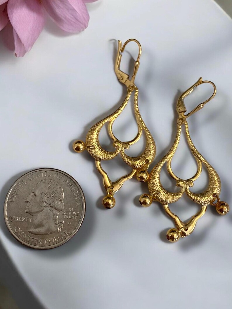May include: A pair of gold-tone dangle earrings with a teardrop shape and intricate detailing. The earrings have a hook closure and are accented with small round beads.